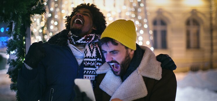 Two friends bundled up in winter clothes celebrating outdoors with holiday lights in the background, looking at a phone.