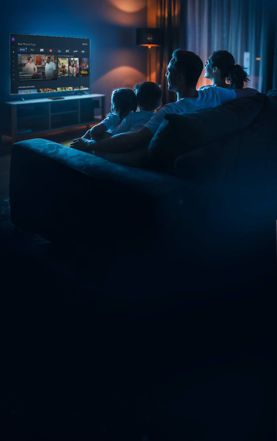 Family sitting together in a dark living room watching DIRECTV on a large TV screen displaying a modern interface with live TV and app content.