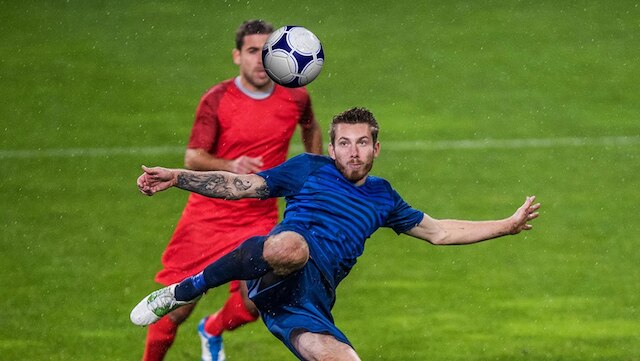 A college soccer player in a red uniform and a player in a blue uniform are competing for the soccer ball on the field